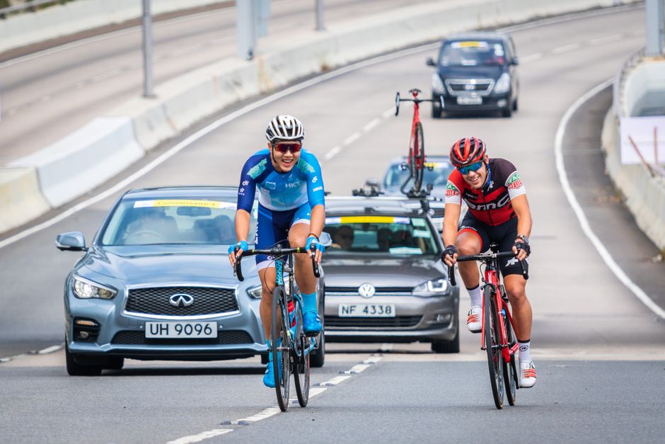 Two cyclists compete in a road race with cars following on an urban highway.